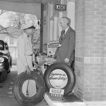 Two men standing outside of the ladie's restroom of a Gulf Service Station next to two tires.