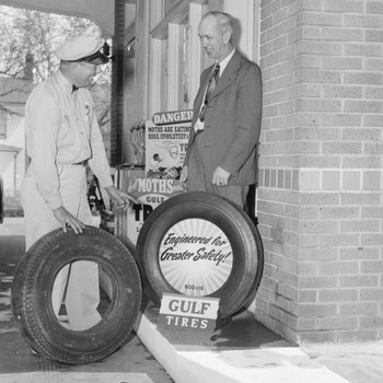 Two men standing outside of the ladie's restroom of a Gulf Service Station next to two tires. 2