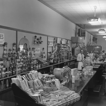Inside of Walter's Restaurant. View of counter with men on both sides looking towards the camera. Candy bars, cans of apple sauce, and other items can be seen for sale behind the counter.