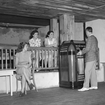 Inside of the Shenandoah Alum Springs Hotel, a group of people sitting and standing, with a man playing a Jukebox. Orkney Springs, Va.