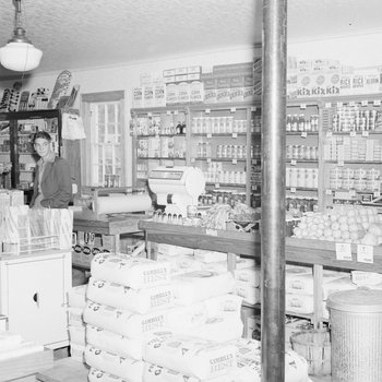 Inside of a store, view of shelving stocked with various food items, and four men standing to the far left.