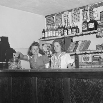 Inside of a store, view of a cash register and two women standing behind the counter.