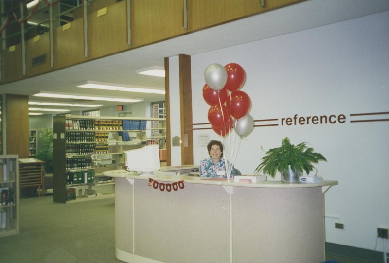 Janet Sinder sits at Reference desk smiling for photo in library, balloons and sign reading, "500,000" hang on desk.