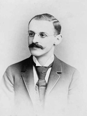 Black and white portrait of Abraham Flexner, a man with small wire-rimmed glasses and a full mustache in a dark suit with a big tie.