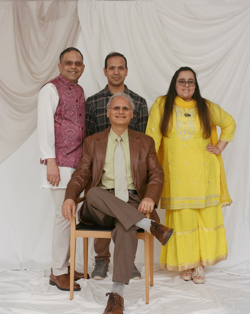 A man of Indian descent is seated in a chair with two men of Indian descent standing behind him. A woman of Indian descent in a yellow outfit is standing to the right of the seated and standing men.