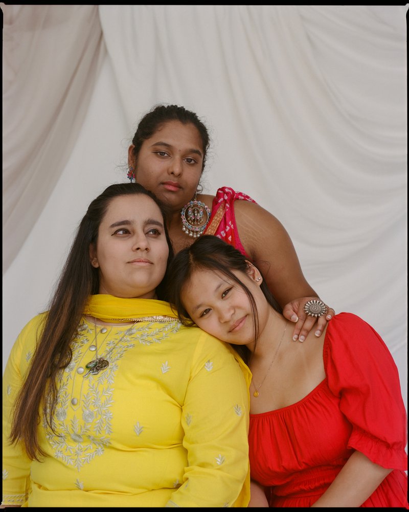 Three women against a white backdrop. Two women are in the front and one woman is behind the two women. The woman on the left is wearing a traditional Indian outfit in yellow. The woman on the right is wearing an orange top. The woman behind them is wearing an orange and white top with large round earrings and a large round ring.