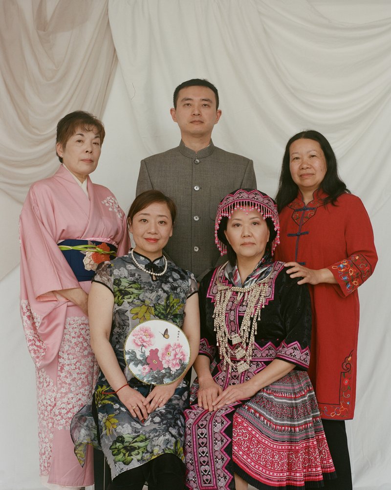 A group of five people, one man and four women, against a white backdrop. All of the people are wearing traditional clothing of Asian countries.