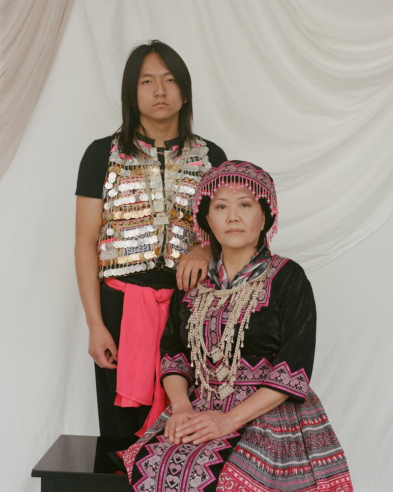 A man and a woman of Hmong descent against a white backdrop. The woman is seated on a bench and the man is standing behind her with his hand on her shoulder. The man and woman are wearing traditional clothing of the Hmong.