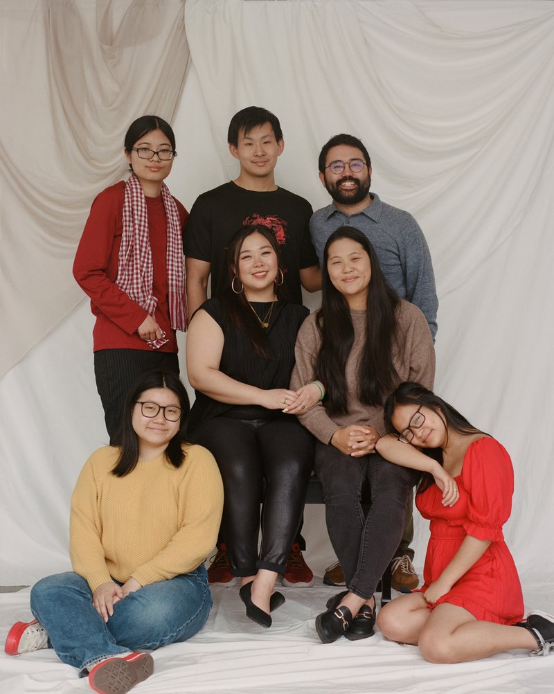 A group of seven people, two men and five women, against a white backdrop. Two women are seated on chairs in the center. One woman is seated on the ground next to the woman in the left chair. One woman is seated on the ground with her head resting on her bicep which is on the seated woman's leg. Two men are standing behind the women seated on chairs. One woman is standing to the left of the two men.