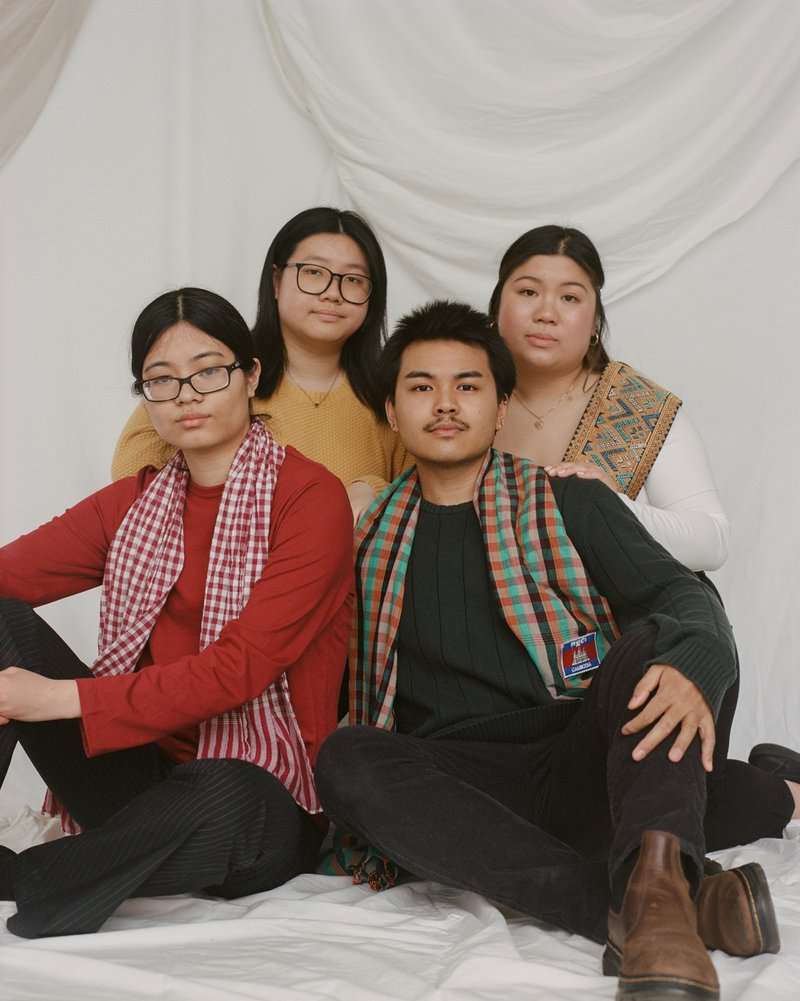 A group of four people, three women and one man seated against a white backdrop. One woman in red top with black pants and a red and white checked scarf is seated on the left. One man wearing all black clothes with an orange, green, and black plaid scarf is seated on the right. There is a woman on the left behind the woman and man in front and one woman on the right behind the seated man and woman.