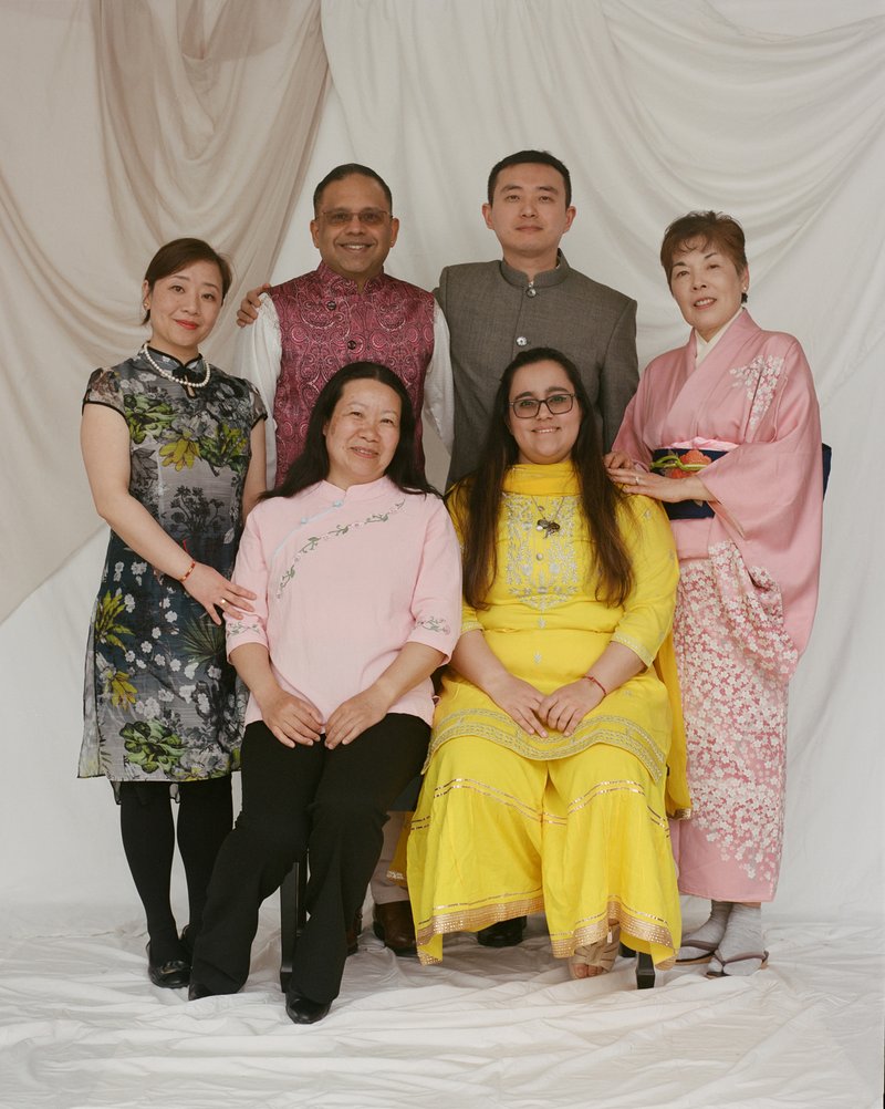 A group of six people, two men and four women against a white backdrop. Two women are seated in the front. Two men are standing behind the seated women. One woman is standing to the left of one standing man and one seated woman. One wooman is standing to the right of one standing man and one seated woman.