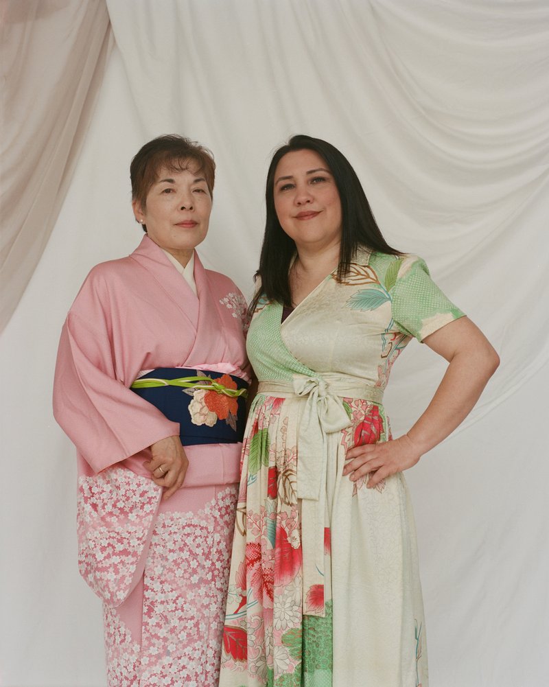 Two women of Japanese descent standing against a white backdrop. The woman on the left is wearing a pink kimono with white blossoms and a black obi with white and coral flowers and a green fabric tie. The woman on the right is wearing a cream colored wrap dress with a fabric tie at the waist and green and coral decoration on the dress.