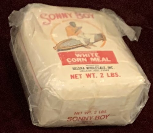 Unopened 2-pound package of "Sonny Boy White Corn Meal. Packed by Helena Wholesale Inc., Helena, Ark." Package features an illustrated Sonny Boy Williamson II, holding a harmonica, sitting on a giant ear of white corn.