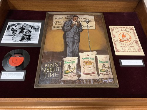 Display case included a photo, an LP record, a painting, and a poster. Promotional photo of Sonny Boy Williamson with Houston Stackhouse and Peck Curtis; Recording of the King Biscuit Radio Hour. Text under LP record: "The only known fully recorded episode of the King Biscuit show was preserved by Chris Strachwitz on this recording he made at KFFA in 1965. (Arhoolie 530 A)"; Painting of Sonny Boy Williamson performing on the King Biscuit Time radio program, signed by “Jimmy Stewart ’90”; Poster, 1987 King Biscuit Blues Festival. Text under poster: "Since 1986, Helena, Arkansas has hosted the annual King Biscuit Blues Festival."