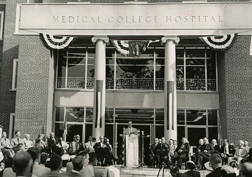 Black and white image of a man speaking at a podium with the Medical College Hospital facade in the background.