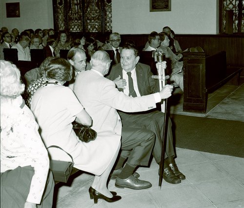 Black and white image of people sitting in pews with the focal point being two men in suits turned toward each other, talking.