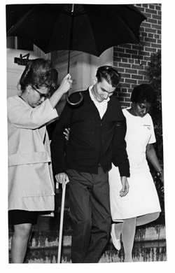 Image of three people walking down the steps. The man in the center has a cane. He is escorted by a woman on the left with an umbrella and a nurse on his right.