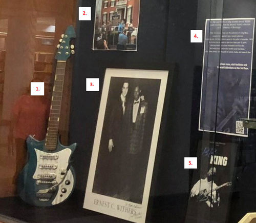 photo of items in display case. includes numbers corresponding to list: guitar, photo of historical marker, framed inscribed photo, timeline of Blues Archive, poster
