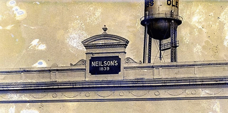 A GIF of two photographs of the top of the department store building with a sign that says "Neilson&#x27;s 1839." One looks older; it is sepia toned and has some discoloration. The other is a modern photograph with the white and navy building standing out against a clear blue sky.