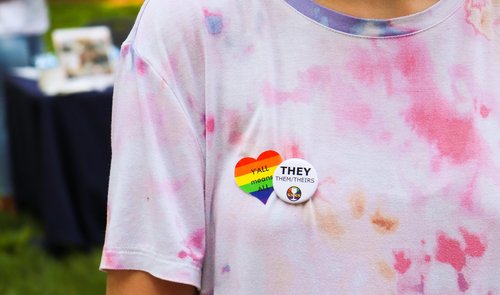 A sticker (Y&#x27;all means all) and a button (They, them, theirs) on a tie-dyed t-shirt