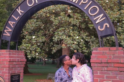 A queer couple kisses under the entrance to the Walk of Champions in the Grove
