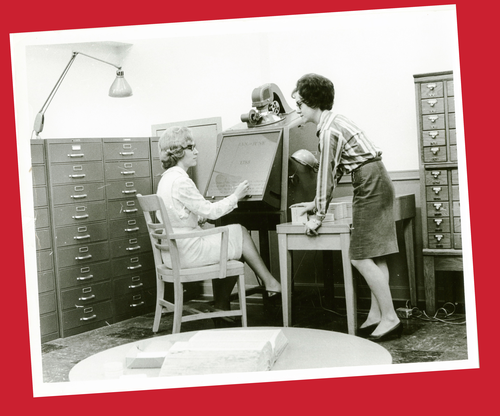 Librarians at a microfilm reader, next to a card catalog.